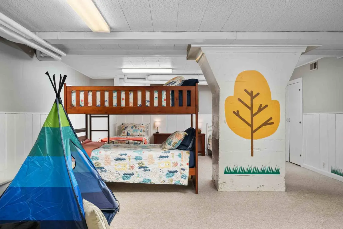 This playful bedroom includes a bunk bed, a small teepee, and a wall mural featuring trees, creating a whimsical and cozy zone for children. The space is ideal for imaginative play, storytelling, and restful sleep.