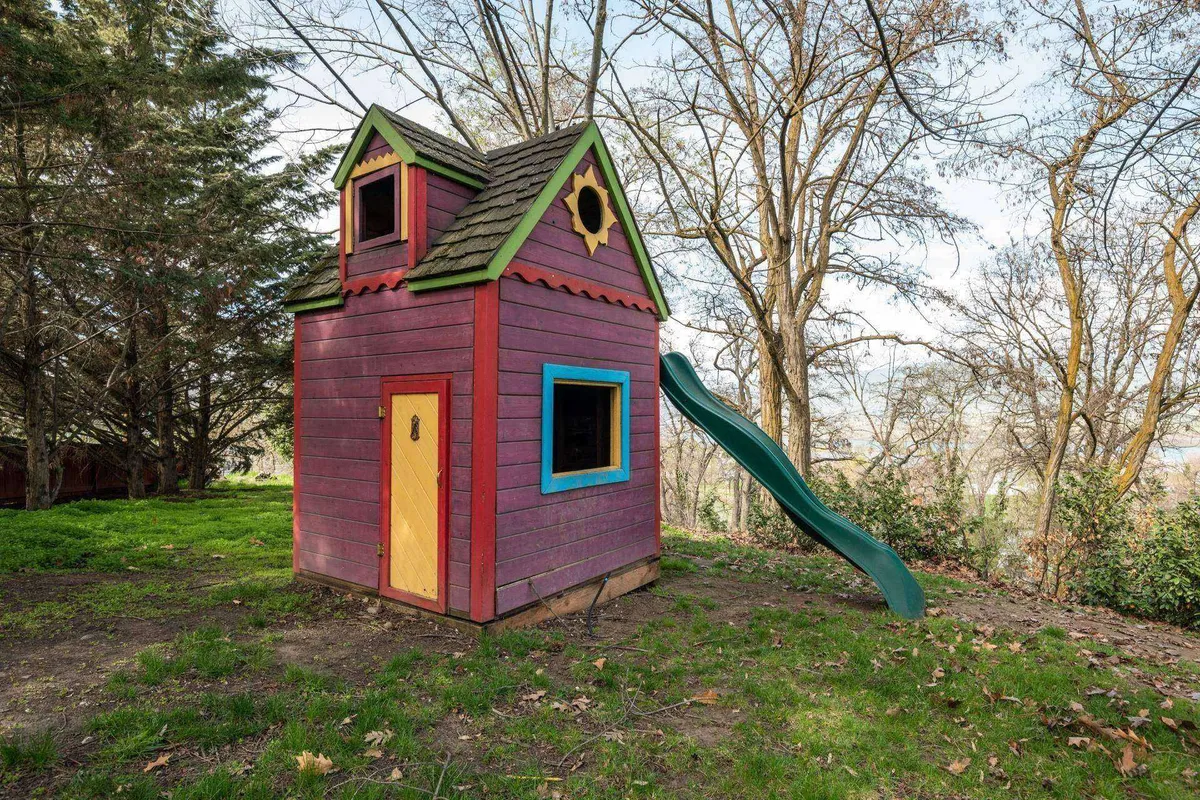 A brightly colored playhouse with a green slide surrounded by mature trees creates a playful, inviting outdoor scene. The area is clearly designed for children's imaginative play and outdoor adventures, tying into the section's emphasis on zones for younger guests.