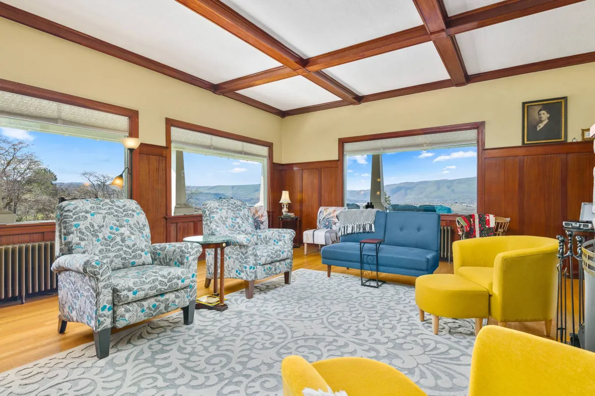 The image showcases a light-filled, inviting living room with plush floral armchairs, a blue sofa, and a vibrant yellow accent chair arranged for conversation. Expansive windows frame sweeping views of the Columbia River Gorge, blending cozy indoor comfort with breathtaking natural beauty. The setting is ideal for relaxing with a morning coffee, gathering for games, or simply soaking in the sunset.