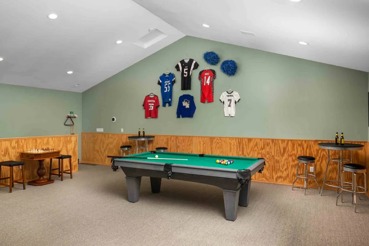 This image captures a lively game room featuring a pool table as the centerpiece, a chess table in the corner, sports jerseys adorning the walls, and bar stools lined up along a counter. The space feels casual and vibrant, encouraging group activities and socialization.