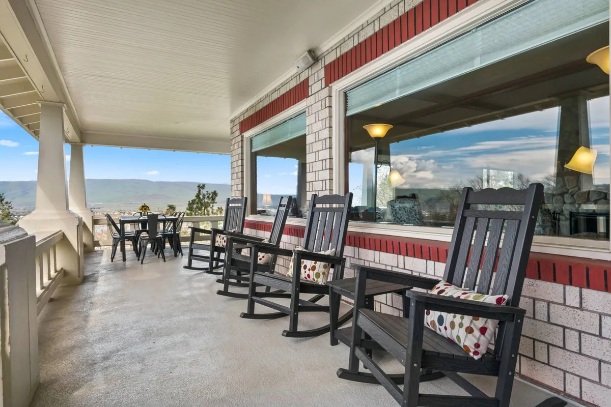 An outdoor patio scene featuring rocking chairs, a dining table, and panoramic mountain views. The space is set up for socializing, dining, and watching the sunset with friends.