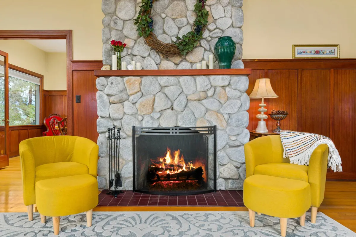 A cozy living area featuring a classic stone fireplace, two bright yellow chairs, a decorative wreath, and a curated vase all set against the warmth of wooden detailing. The room is arranged for intimate gatherings or quiet relaxation, reflecting a harmonious blend of heritage and comfort.