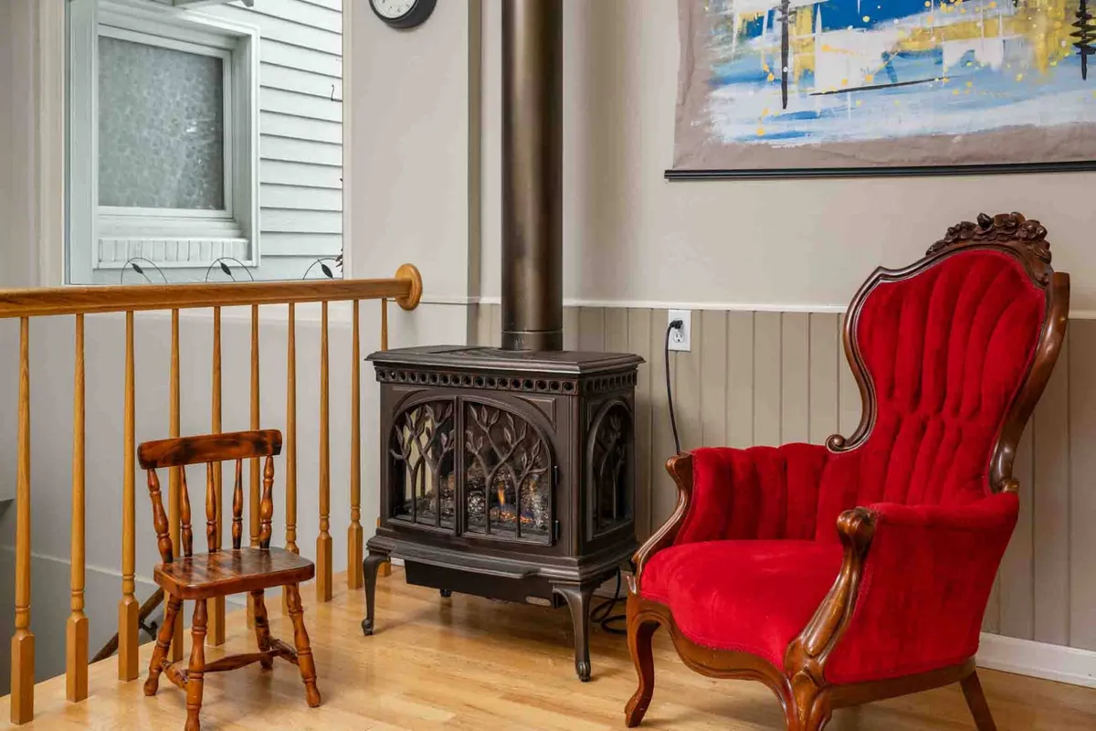 A cozy corner featuring a red armchair and a wooden stool beside a stove, with thoughtfully curated wall art. The setting exudes warmth and invites guests to unwind, read, or share heartfelt conversations in a peaceful environment.