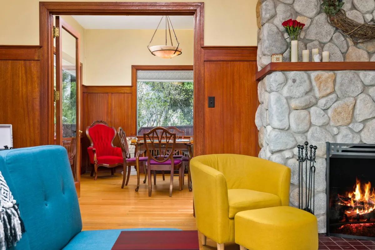 An inviting living and dining space with colorful, eclectic furniture and a fireplace. Large windows let in abundant natural light, offering views of lush greenery outside. The setting is ideal for socializing, reading, or enjoying meals, with touches of local art enhancing the heritage feel.