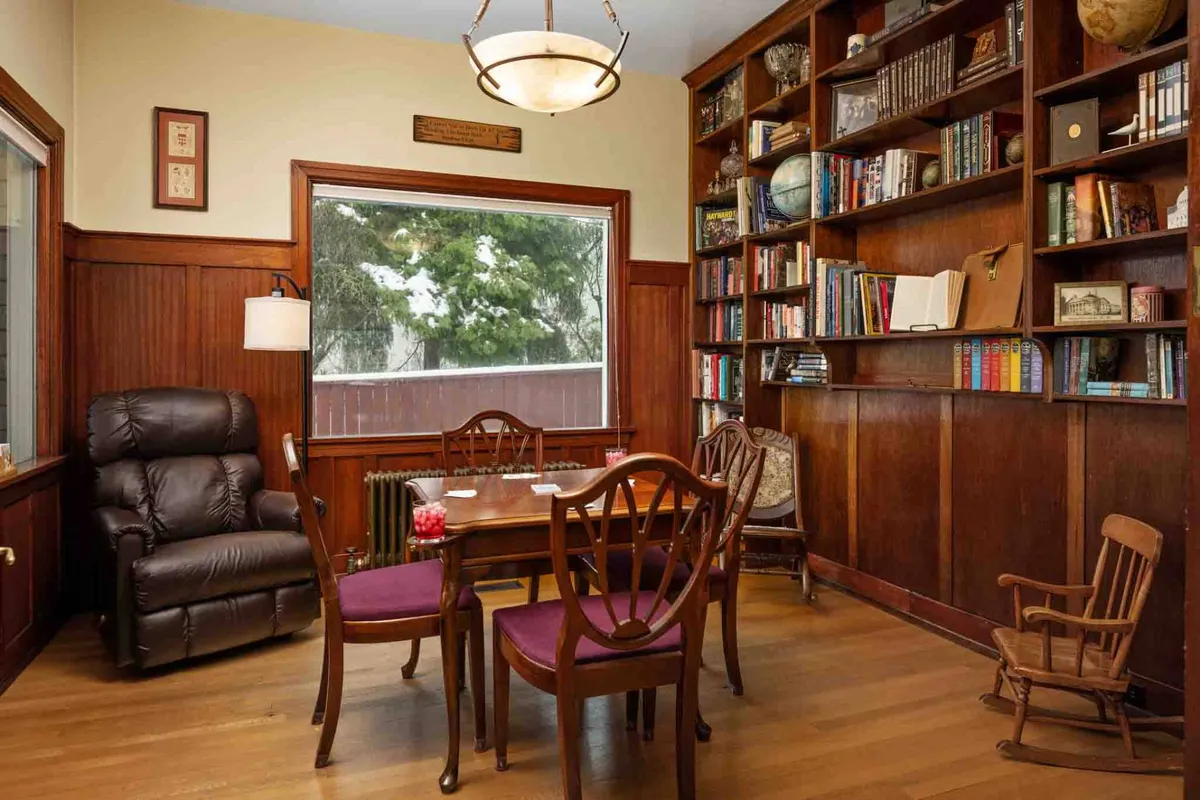 A cozy library nook with a wall of bookshelves, a wooden table and chairs, a comfortable armchair, and a window that invites in natural light and offers a tranquil view. The space feels intimate and perfect for reading, playing games, or enjoying heartfelt conversation.